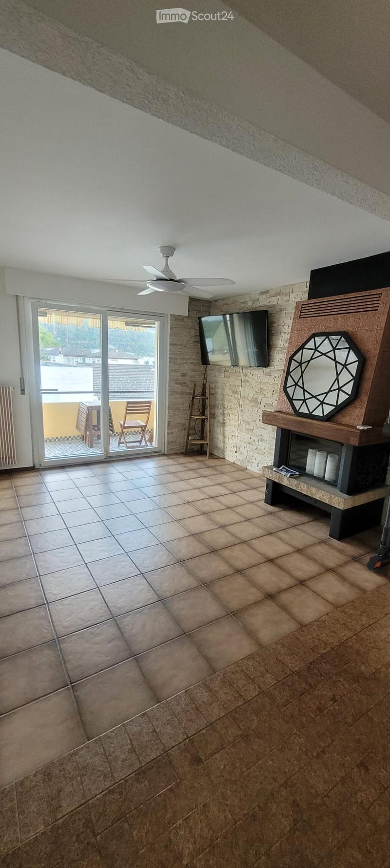 Living room with tiled floor, white ceiling fan, television mounted on wall, decorative mirror on wooden stand, and sliding glass door leading to balcony with outdoor furniture.