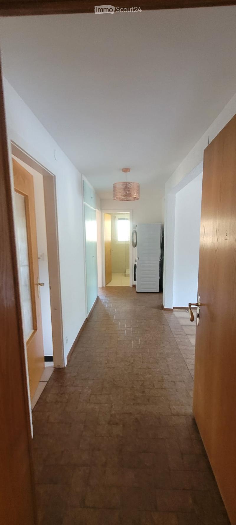 A long, narrow hallway with tiled flooring, white walls, and wooden doors on both sides. A hanging light fixture is visible in the center, and a glimpse of a room with a refrigerator can be seen at the end.