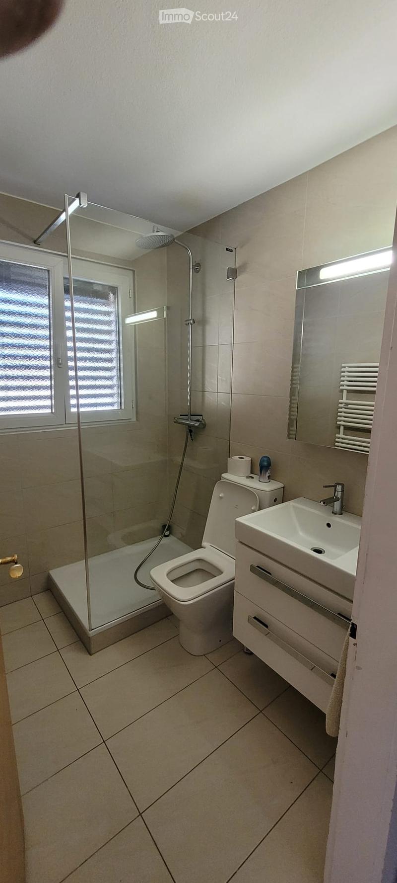 Modern bathroom with beige tiles, a glass-enclosed shower, a toilet, a sink with a mirror, and a towel rack.