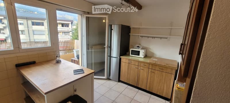 Modern kitchen with wooden cabinets, stainless steel refrigerator, microwave, and a wooden table with a tablet on it. Large windows and a sliding glass door lead to a balcony.
