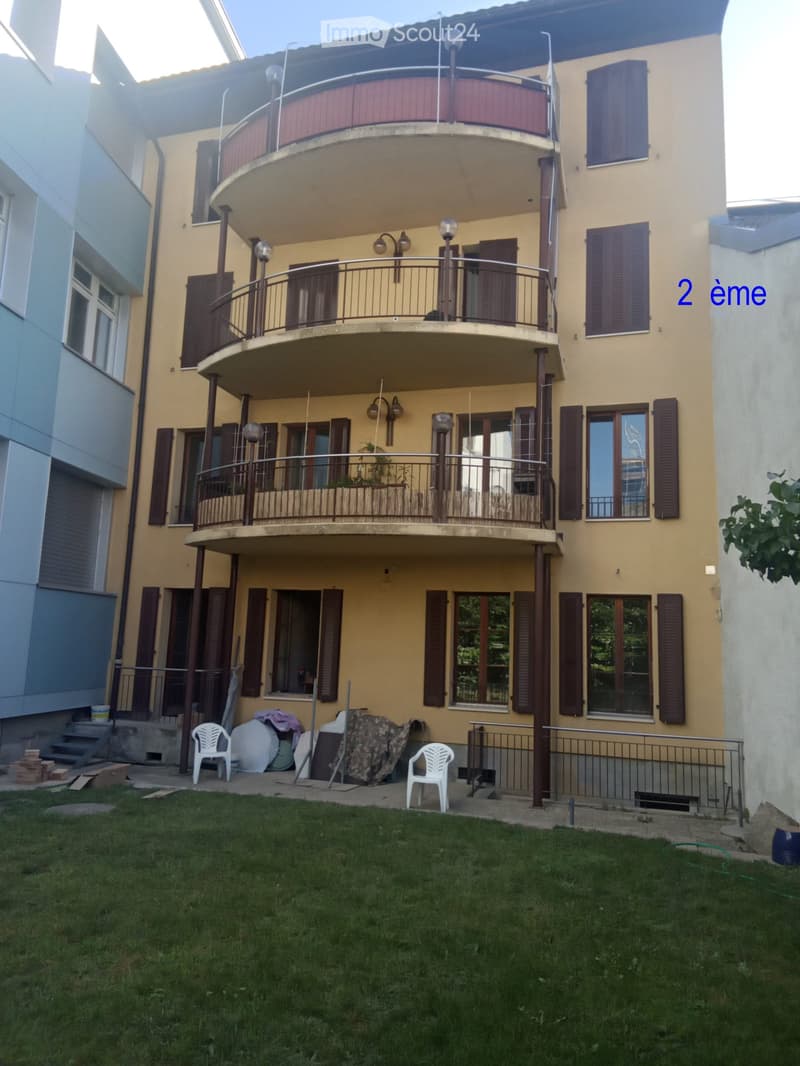 Two-story house with yellow facade, multiple windows with shutters, balconies on both floors, and a small grassy yard with a few plastic chairs and construction materials.