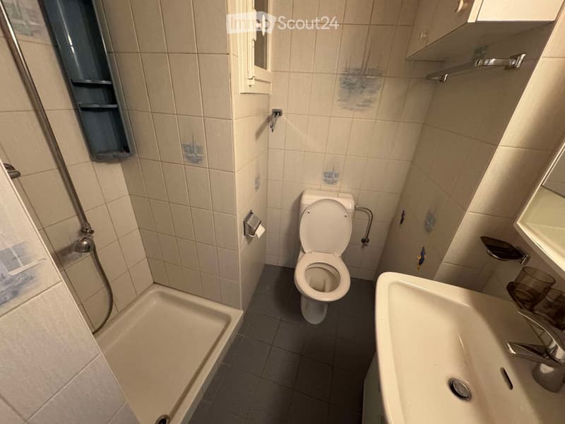 A small bathroom with beige tiled walls and dark tiled floor, featuring a toilet, sink, bathtub, and mirror.