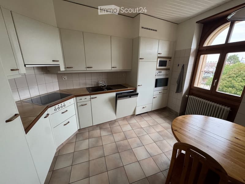 Modern kitchen with white cabinets, tiled walls, and a wooden dining table with a chair. Appliances include an oven, microwave, and dishwasher.