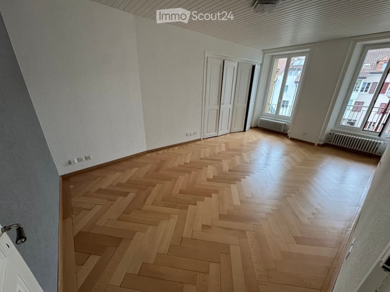 Empty room with wooden herringbone flooring, white walls, and large windows with radiators beneath them. The room has a slanted ceiling and a door leading to another space.