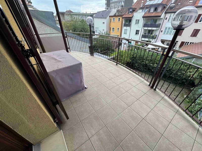 Balcony with tiled floor, metal railing, and a covered table, overlooking a cityscape with buildings and greenery.