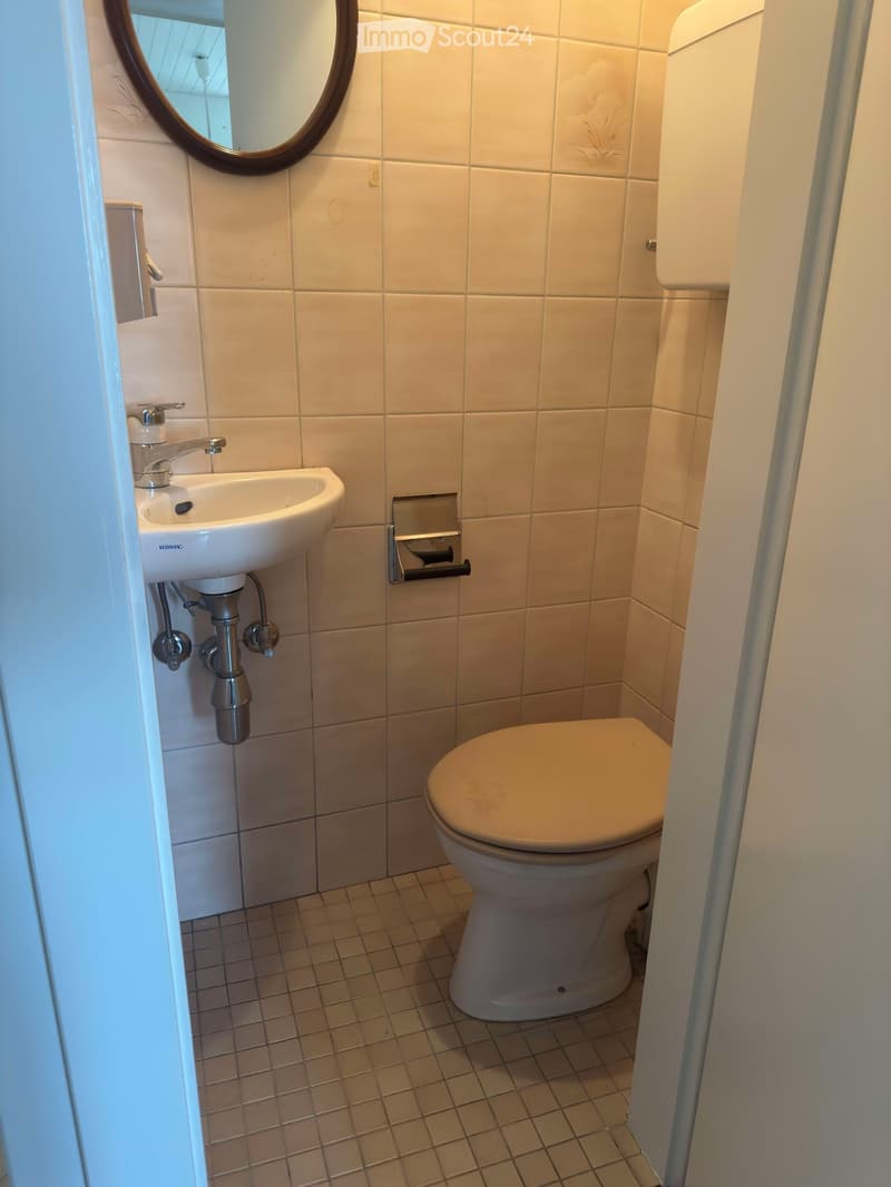 Small bathroom with white tiled walls and floor, featuring a toilet, sink with a chrome faucet, and a round mirror above the sink.