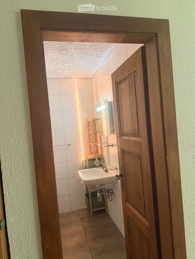 A small bathroom with white tiled walls, a wooden door frame, a sink with a modern faucet, a mirror with built-in lighting, and a wooden shelf unit.