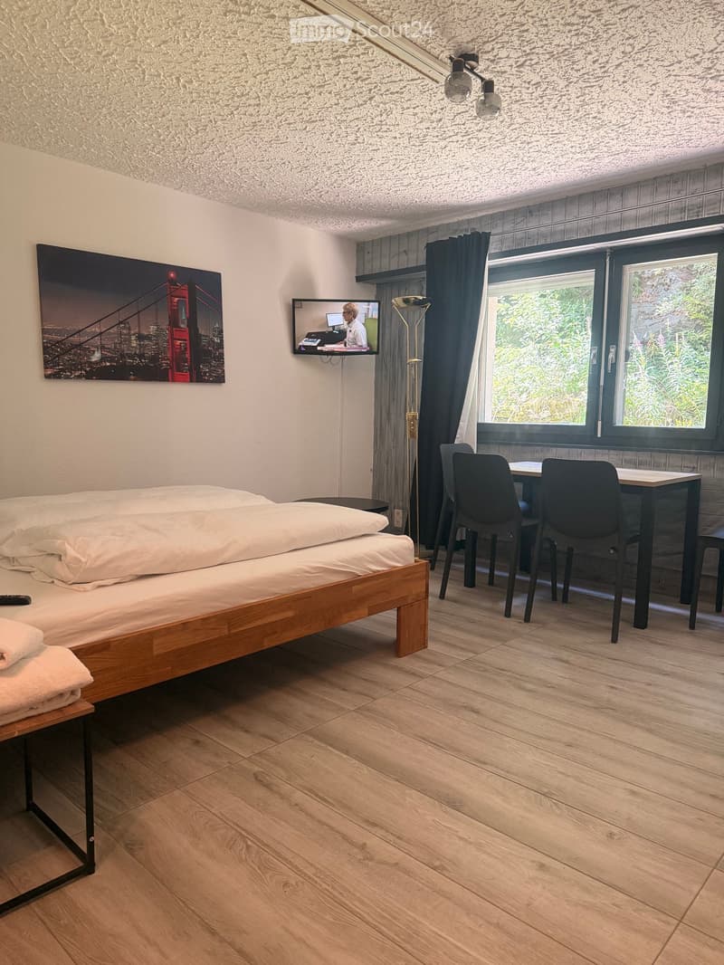 Cozy bedroom with a wooden bed, a small table with chairs, a mounted TV screen, and a large window with a view of greenery.