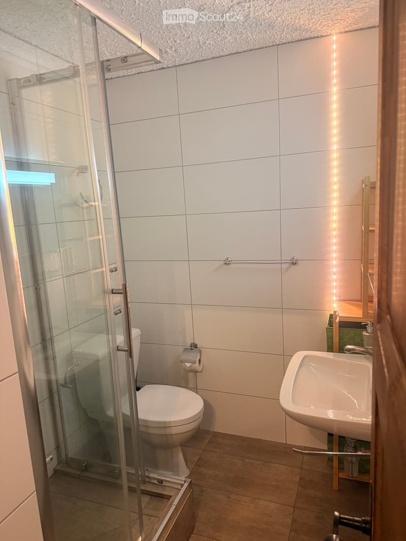 Modern bathroom with white tiles, a glass-enclosed shower, a toilet, a sink, and a mirror. The space is well-lit with a sleek design.