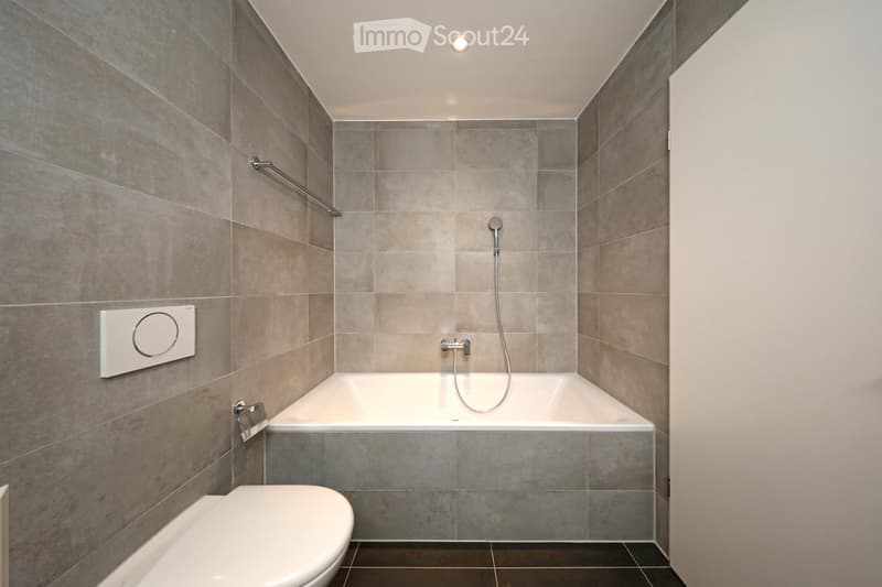 Modern bathroom with a bathtub, toilet, and showerhead, featuring grey tiled walls and a sleek design.
