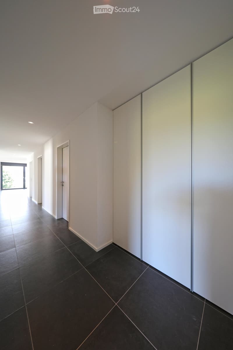 Empty hallway with dark tiled floor, white walls, and recessed lighting. Features a large window on the left and a closed door on the right.