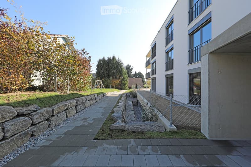 Modern residential complex with a paved pathway, stone walls, and a small garden area with trees and shrubs.
