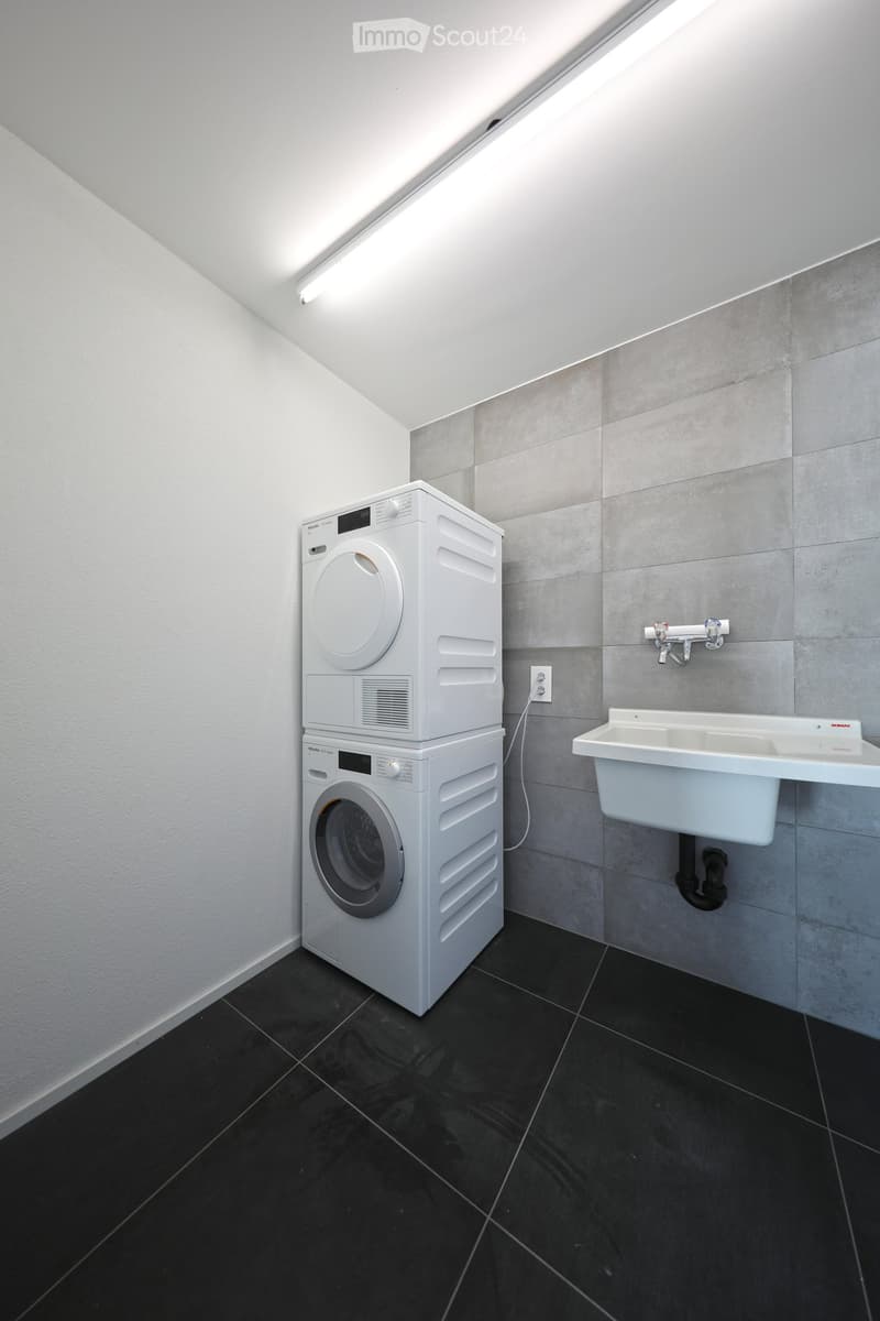 Modern bathroom with a stacked washing machine and dryer, a white sink, and grey tiled walls.