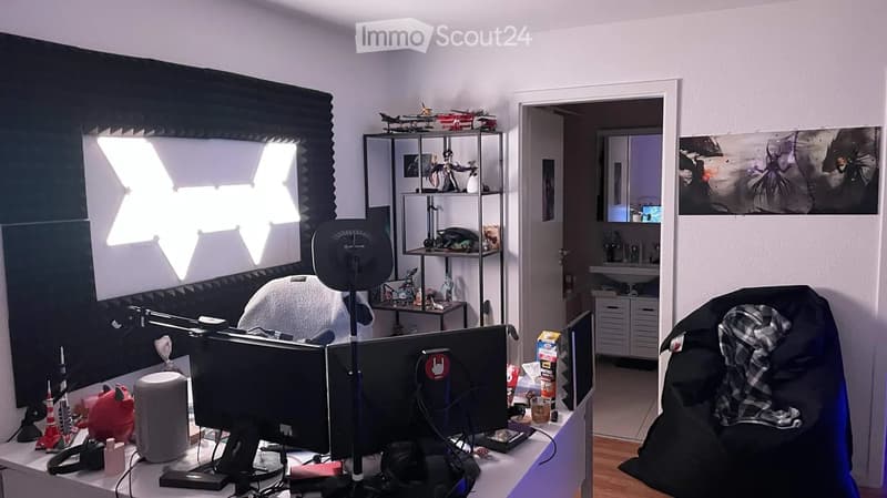A well-lit room with a desk setup, featuring a chair, table, screen, shelf, and various items including a fridge, sink, and toys. A doorway leads to another room with a sink and cabinets.