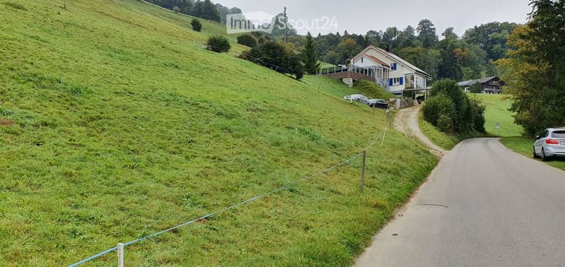 A rural road winds through a lush green hillside with a house on the slope and a few parked cars.