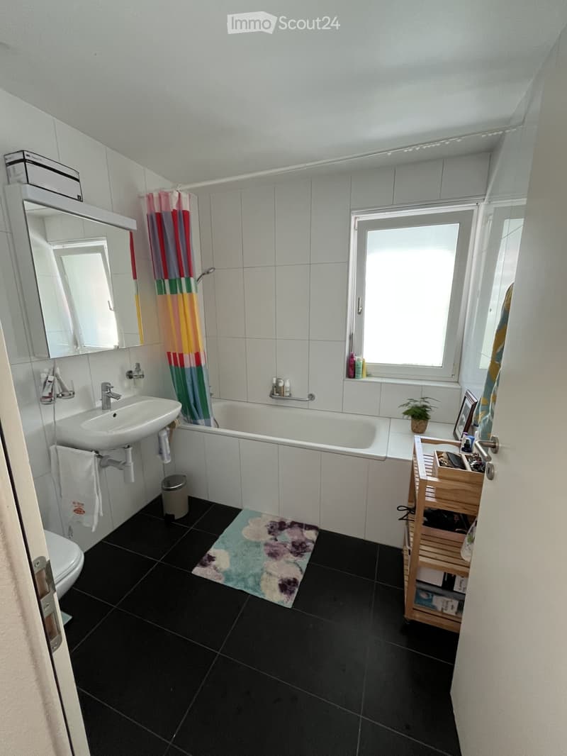 Bathroom with black tiled floor, white tiled walls, sink, bathtub, mirror, toilet, towel, colorful shower curtain, small plant, and a wooden stool.