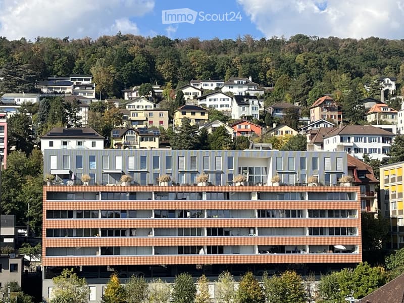 Modern multi-story apartment building with balconies and large windows, surrounded by greenery and other residential structures.