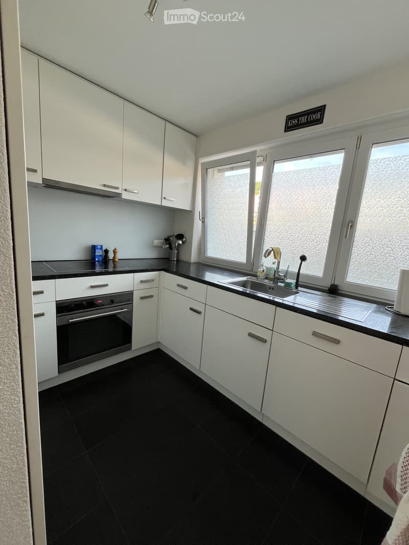 Modern kitchen with white cabinets, black countertops, and a black tiled floor. Features an oven, sink, and various small appliances.