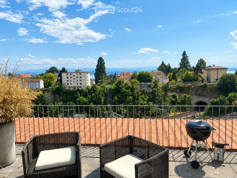 Balcony with wicker chairs, cushions, and a grill, overlooking a cityscape with trees and buildings.