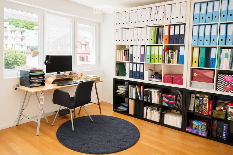 A well-organized home office with a wooden desk, computer setup, ergonomic chair, large bookshelf filled with books and file folders, and a round dark rug on the floor.