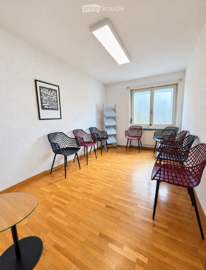 Bright room with wooden flooring, several modern chairs, a round glass table, and a small shelf against the wall.