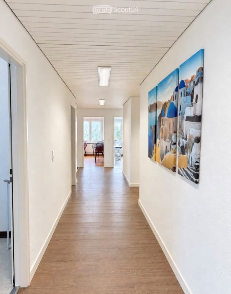Clean, modern hallway with light wooden flooring, white walls, and recessed lighting. Two large paintings are displayed on the right wall, and a partially visible room with chairs and a table is seen at the end of the corridor.