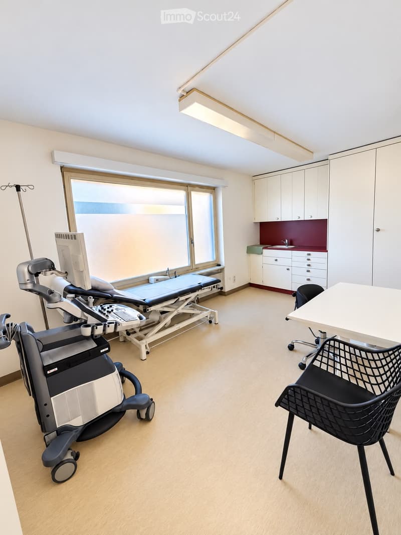 A well-lit medical examination room with a large window, a medical examination bed, a table, a chair, a cabinet, a sink, and medical equipment.
