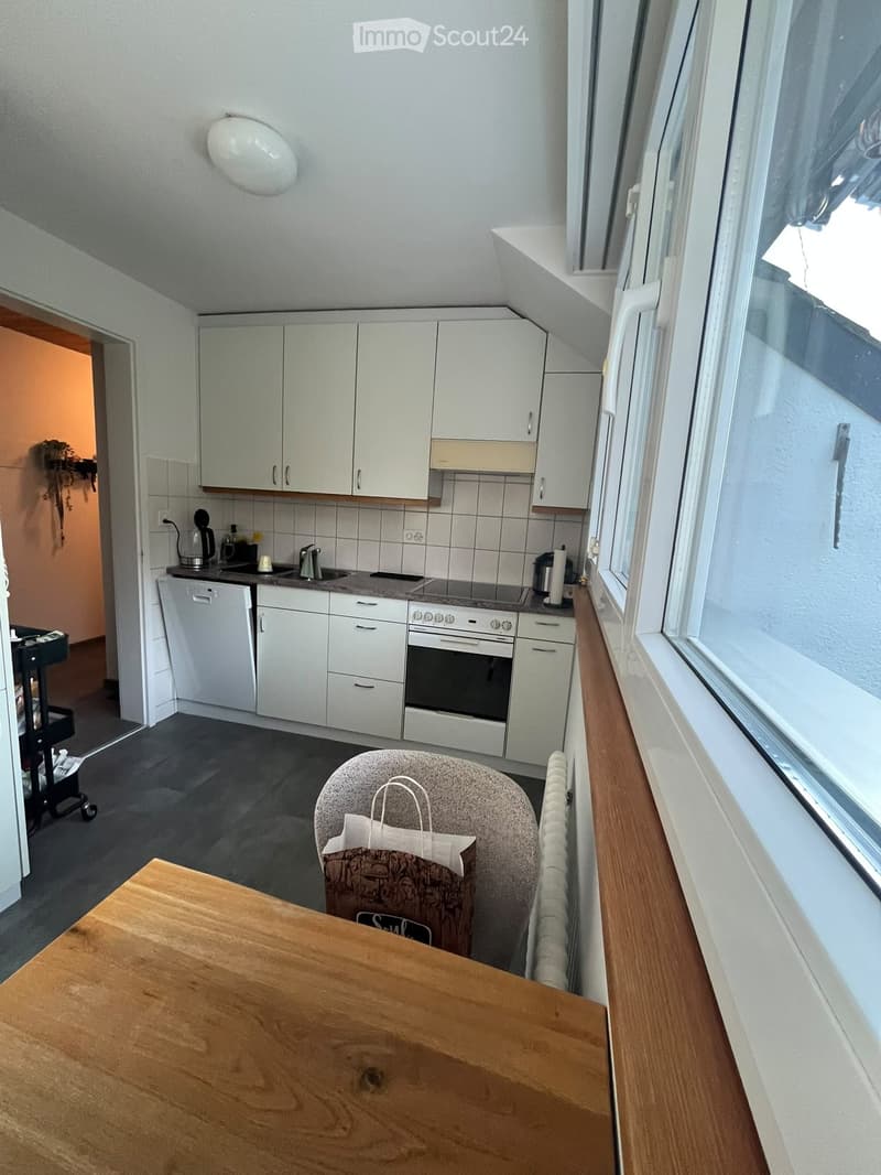 A compact kitchen with white cabinets, a wooden table, a chair, and various appliances including an oven, dishwasher, and sink.