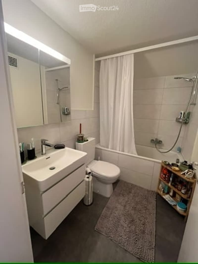 Modern bathroom with a white sink, mirror, toilet, bathtub, and shower curtain. Includes a small shelf with toiletries and a gray bath mat.