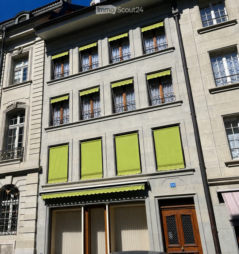 A multi-story building with light-colored stone facade, multiple windows with green awnings, and ornate metal railings on balconies.