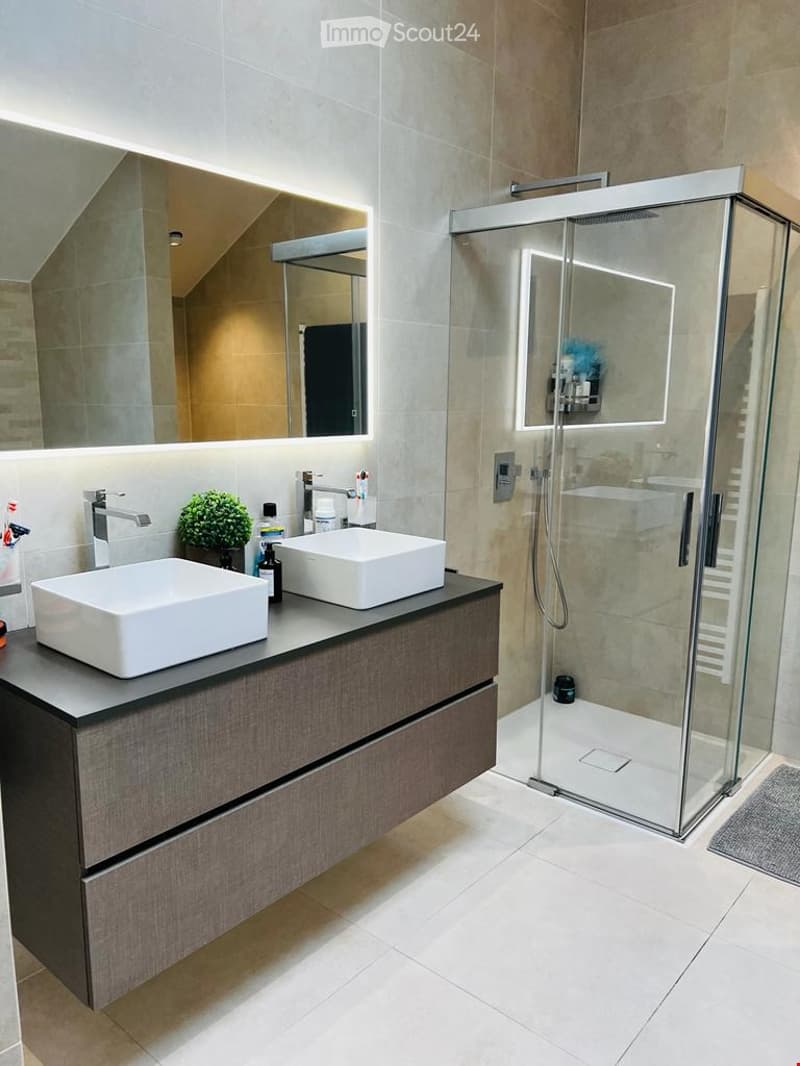 Modern bathroom with two white sinks, a dark wooden vanity, a large mirror with LED lighting, and a glass-enclosed shower with a built-in shelf.