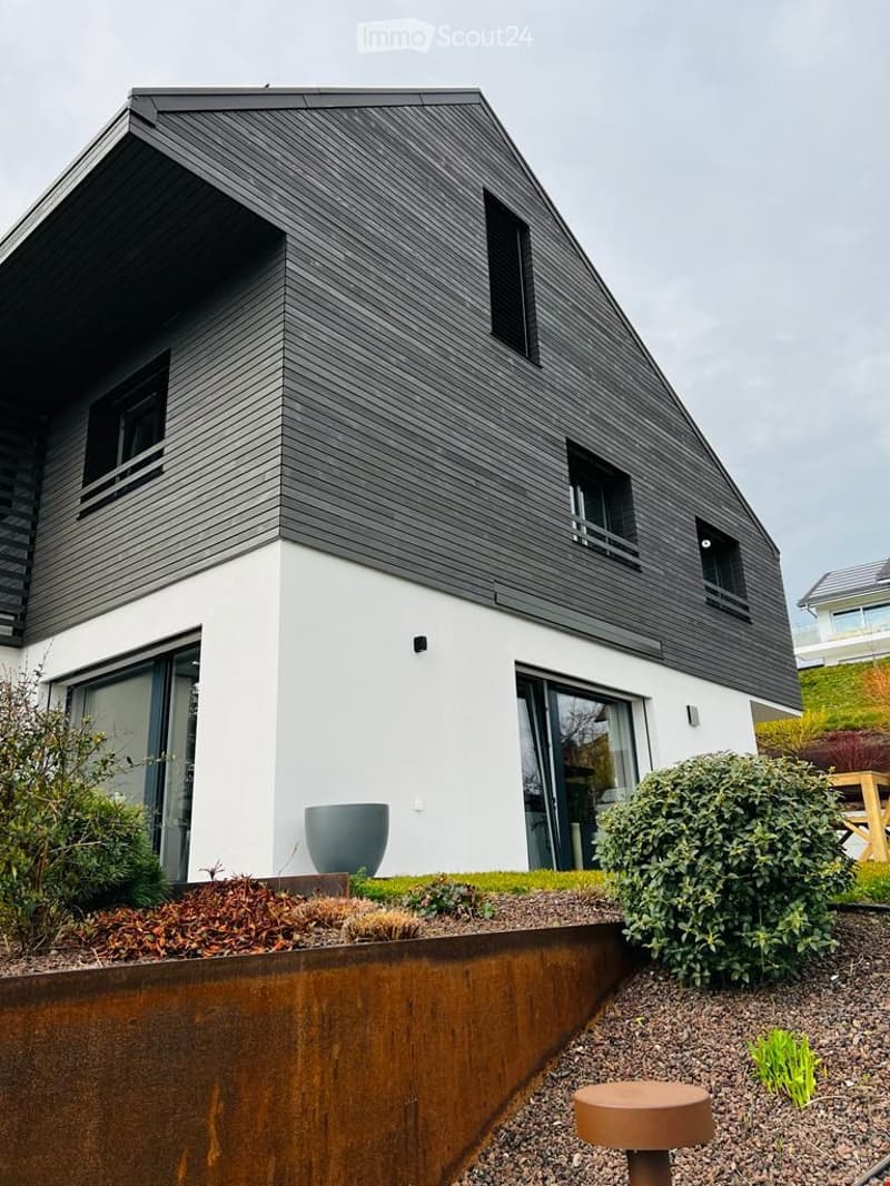 Modern two-story house with dark grey shingled roof and white walls, featuring large windows and a raised ground floor.