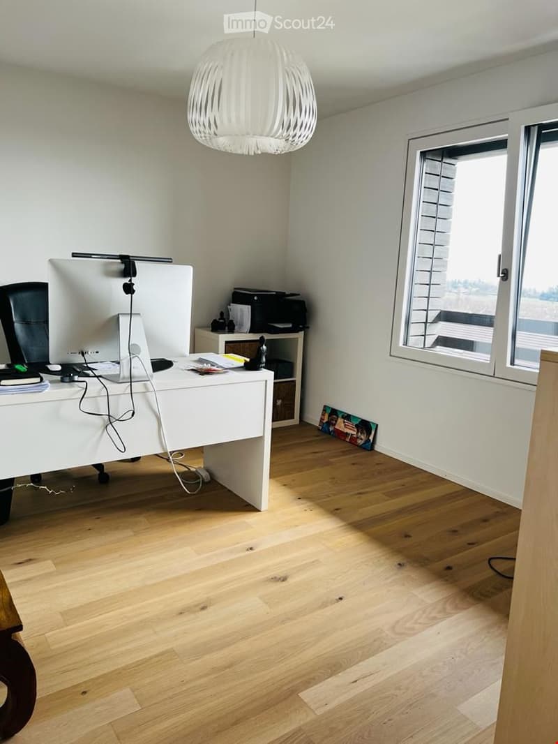 Modern office space with light wooden flooring, white walls, a large window, a white desk with a computer monitor, chair, and printer, and a hanging white lamp.