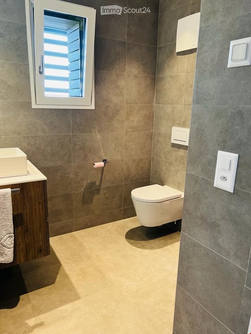 Modern bathroom with beige tiles, a toilet, a sink with a wooden cabinet, and a towel hanging on the side.