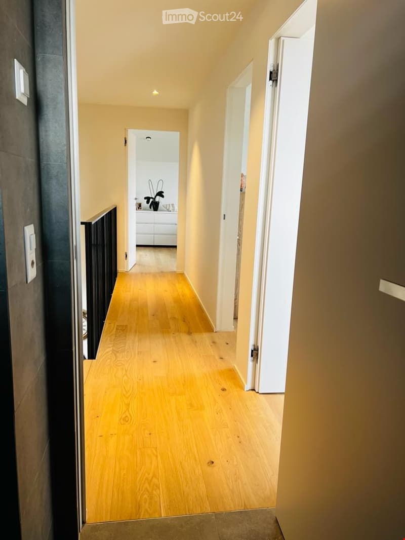 A narrow hallway with light wooden flooring, white walls, and recessed lighting. A staircase leads upwards, and an open doorway reveals a glimpse of a room with a white cabinet and a plant.