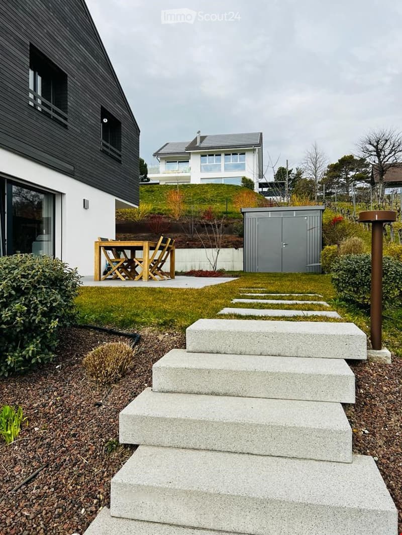 Modern garden area with a raised concrete staircase leading up to a wooden patio set, small lawn, shrubs, and a modern house in the background.