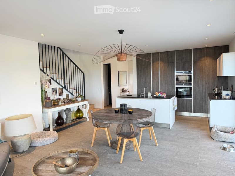 Open-plan living and dining area with a modern kitchen, round dining table, chairs, decorative vases, and a staircase leading upstairs.