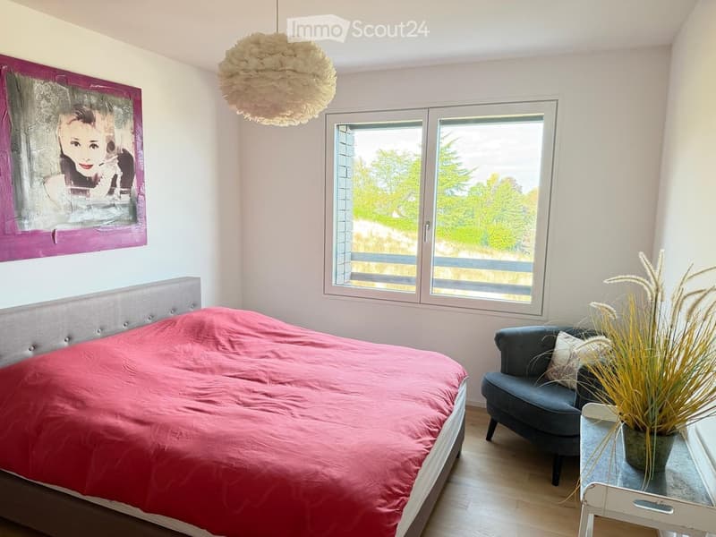 Bright bedroom with a double bed, grey headboard, pink duvet, grey sofa, small table with a potted plant, and a large window with a scenic view.