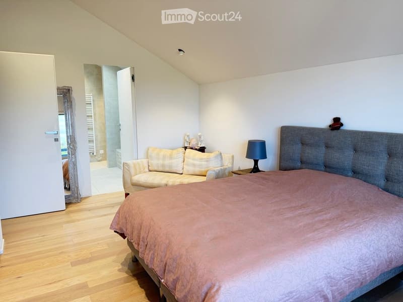 Cozy bedroom with a large bed, a small sofa, a table lamp, and a mirror. The room features wooden flooring and has an open doorway leading to a bathroom.