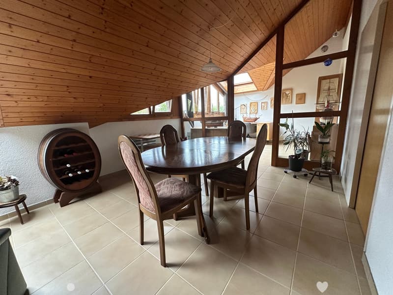 Dining area with a round wooden table, six chairs, a wine rack, and potted plants. The room has a slanted wooden ceiling and large windows.