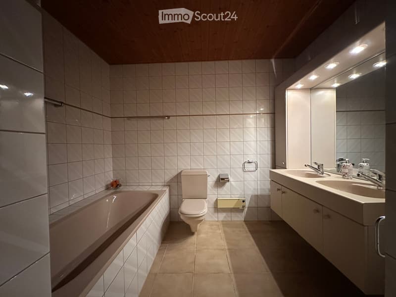 Modern bathroom with a bathtub, toilet, sink, and mirror. Features tiled walls and a wooden ceiling.