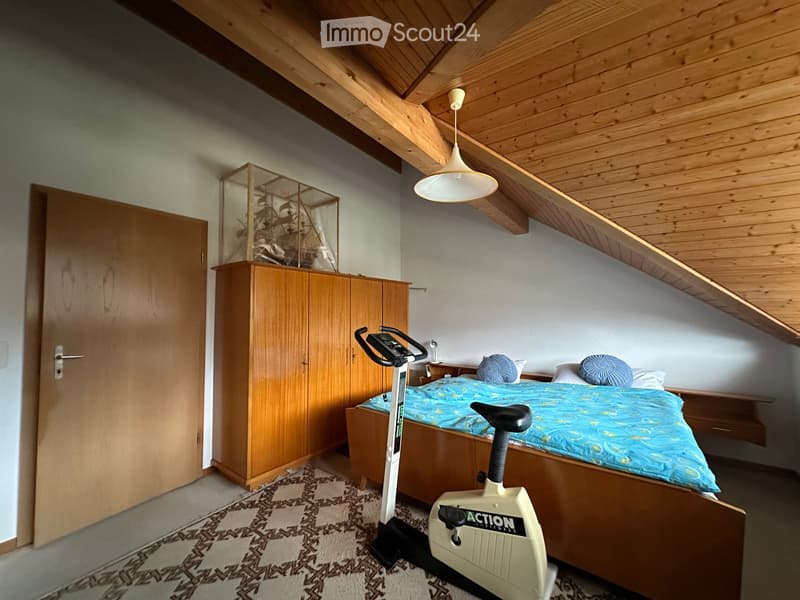 Cozy bedroom with wooden slanted ceiling, bed with blue bedding, exercise bike, wardrobe, nightstand, and mirror.