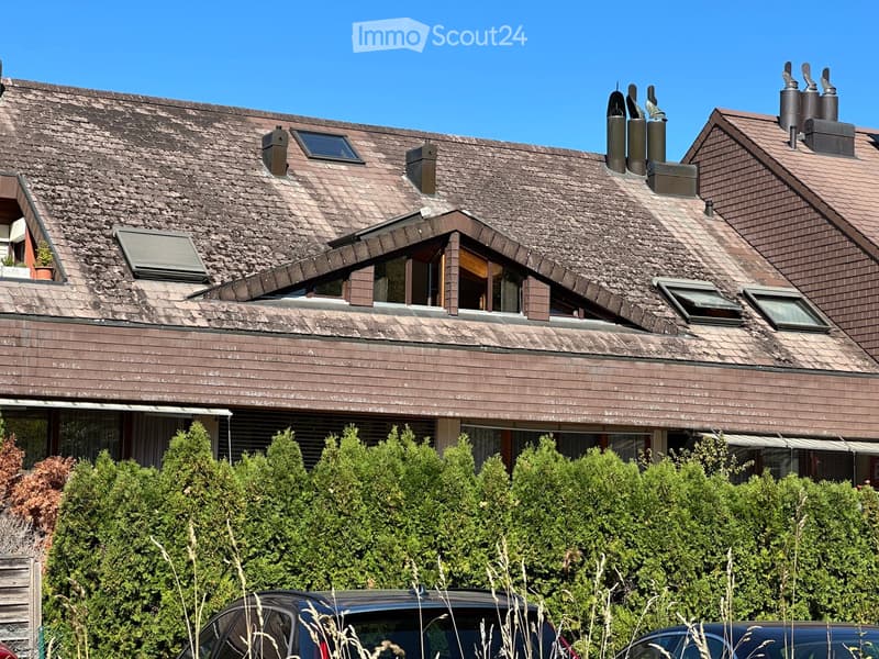 Two-story house with a brown tiled roof, multiple chimneys, dormer windows, and a well-maintained garden with shrubs and a parked car.