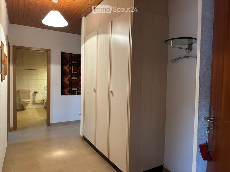 A narrow hallway with tiled flooring, white walls, and a wooden ceiling. A coat rack is mounted on the right wall, and a bathroom is visible to the left with a toilet and tiled walls.
