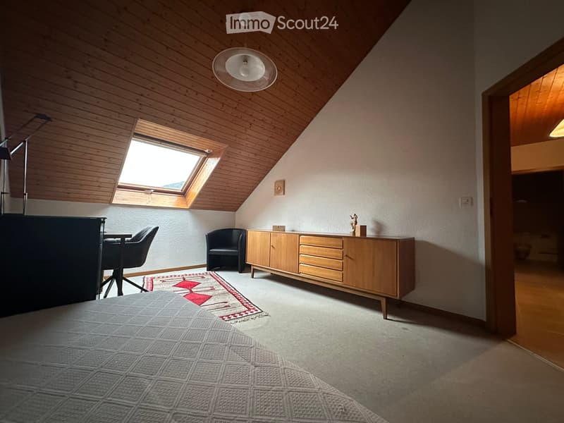 Cozy attic bedroom with wooden slanted ceilings, skylight window, and modern furniture including a bed, chairs, a table, and a wooden cabinet.