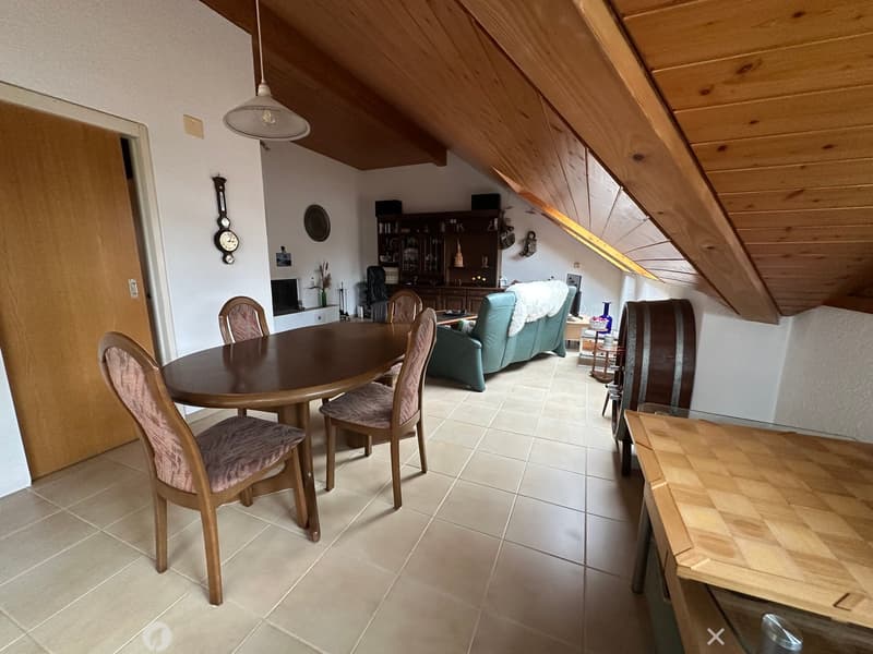 Cozy attic room with slanted wooden ceilings, dining table with chairs, green sofa, bookshelf, barrel, and clock on the wall.