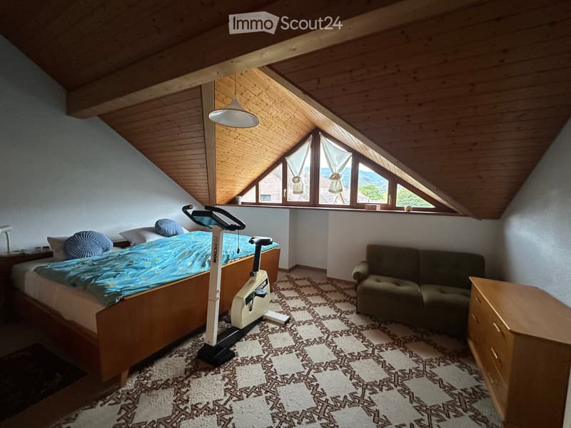 Cozy attic bedroom with wooden beams, large windows, a bed with blue bedding, a sofa, an exercise bike, and a nightstand with a lamp.