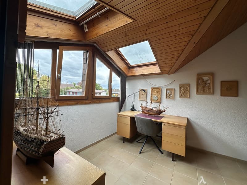 Brightly lit room with wooden ceiling, large windows, model ship on display, desk with chair, lamp, and decorative picture frames on the wall.