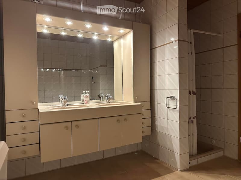 Modern bathroom with double sink, large mirror with integrated lighting, tiled walls, and a separate shower area.