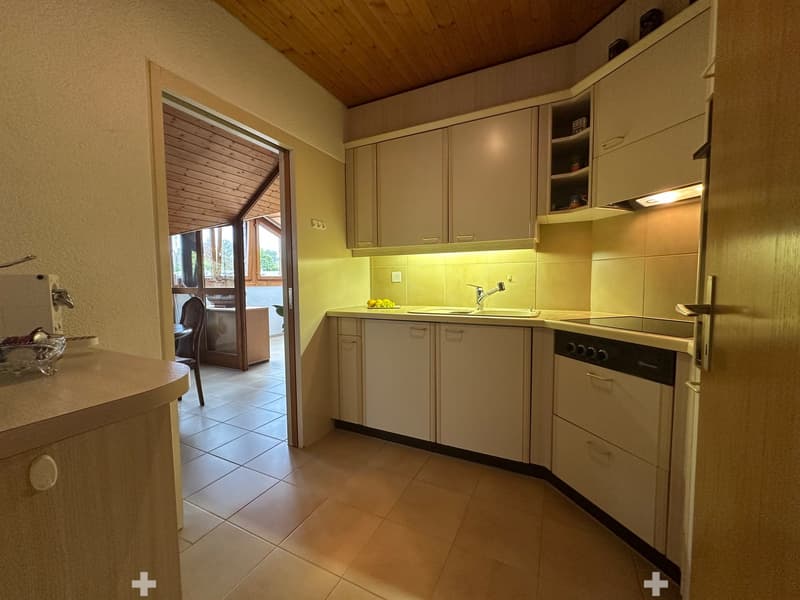 Modern kitchen with light-colored cabinets, a sink, a dishwasher, and a small table with a chair in the adjacent area. The kitchen has a tiled floor and a view to a balcony or terrace through an open doorway.